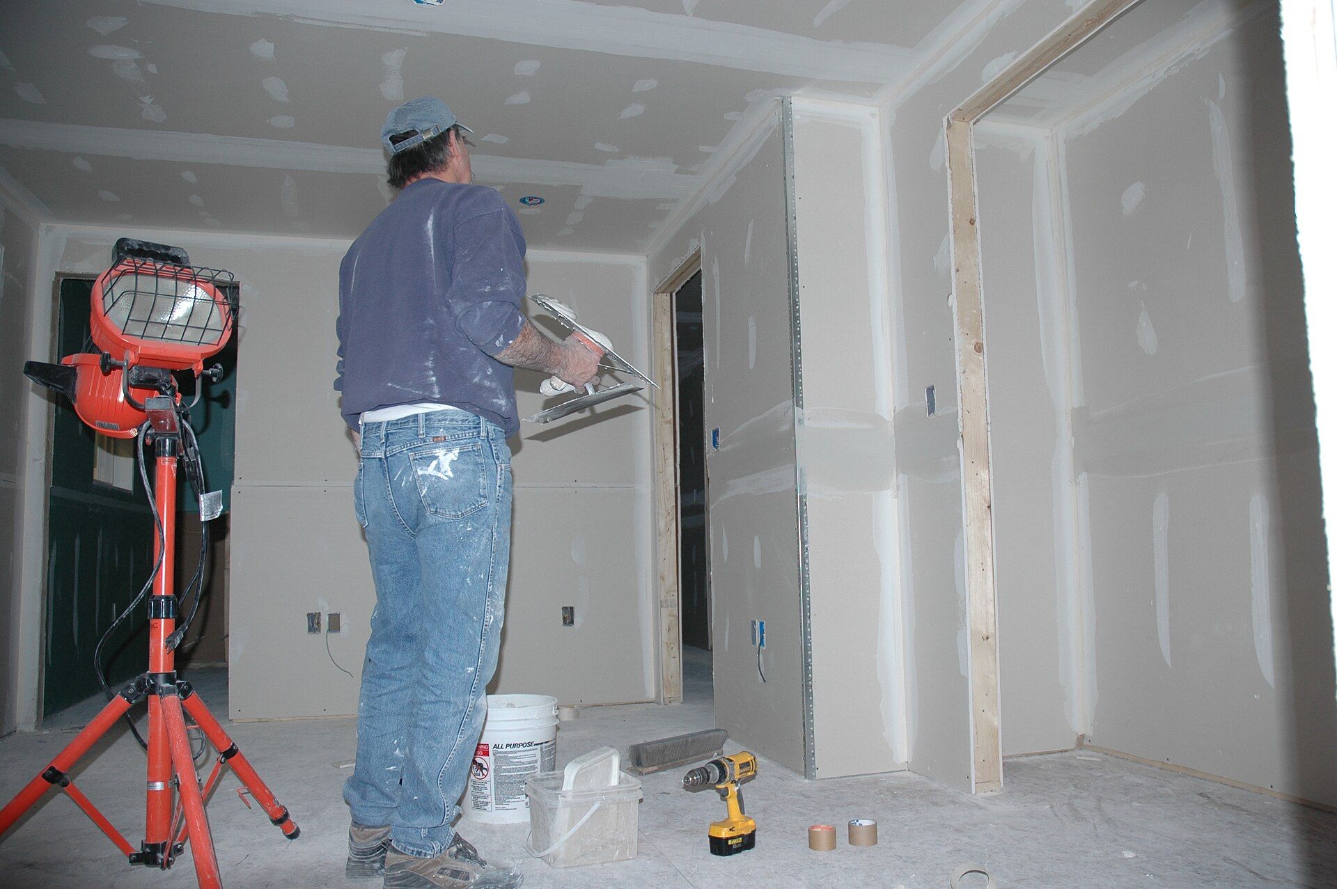 Freshly taped drywall walls in a bare room under a work light, a worker finishing a seam