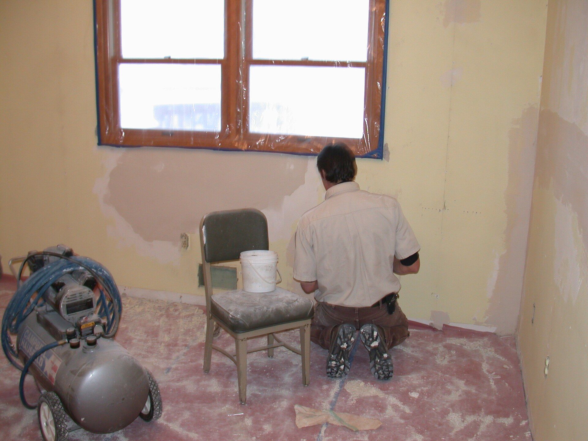 Man patching an old plastered wall under a window, a compressor beside him