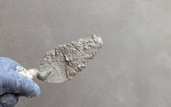 Gloved hand holding a trowel flat against a gray plaster wall